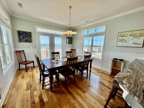 A dining room with a wooden table and chairs, large windows, and a view of the ocean.