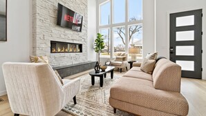 A modern living room with a fireplace, a large TV mounted on a stone wall, and a large window providing natural light.
