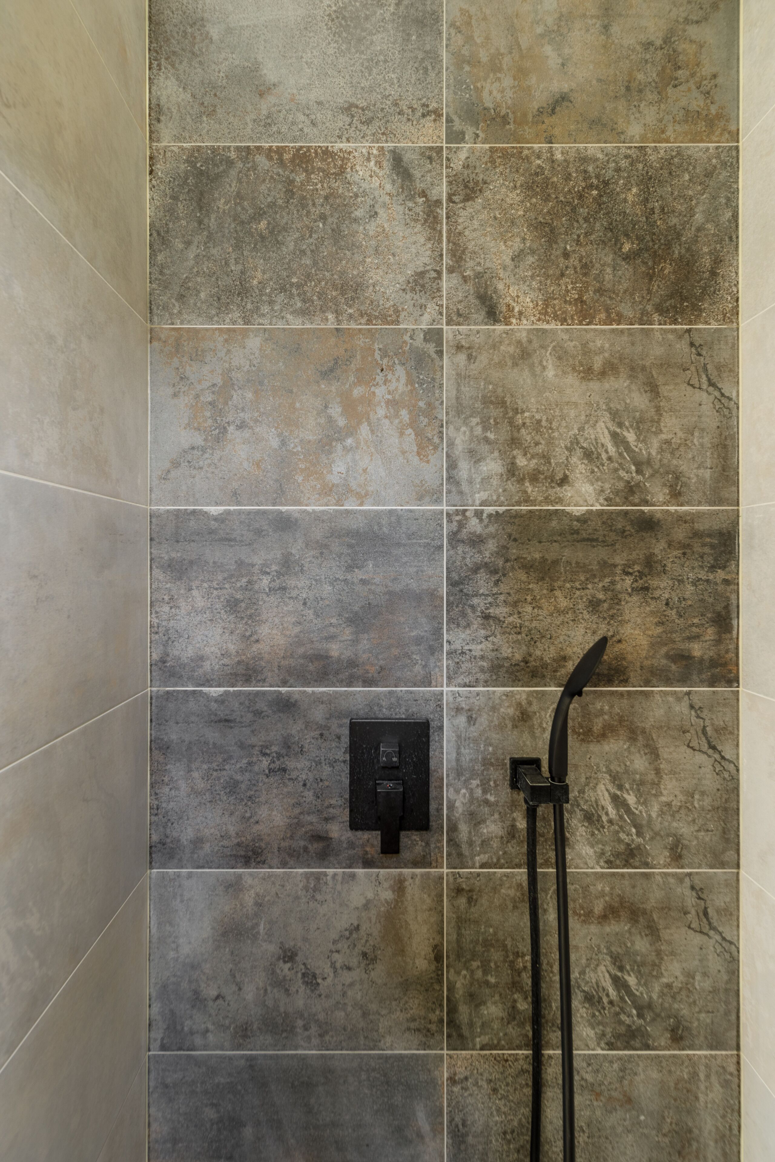 A tiled shower wall with a control panel and a handheld showerhead.