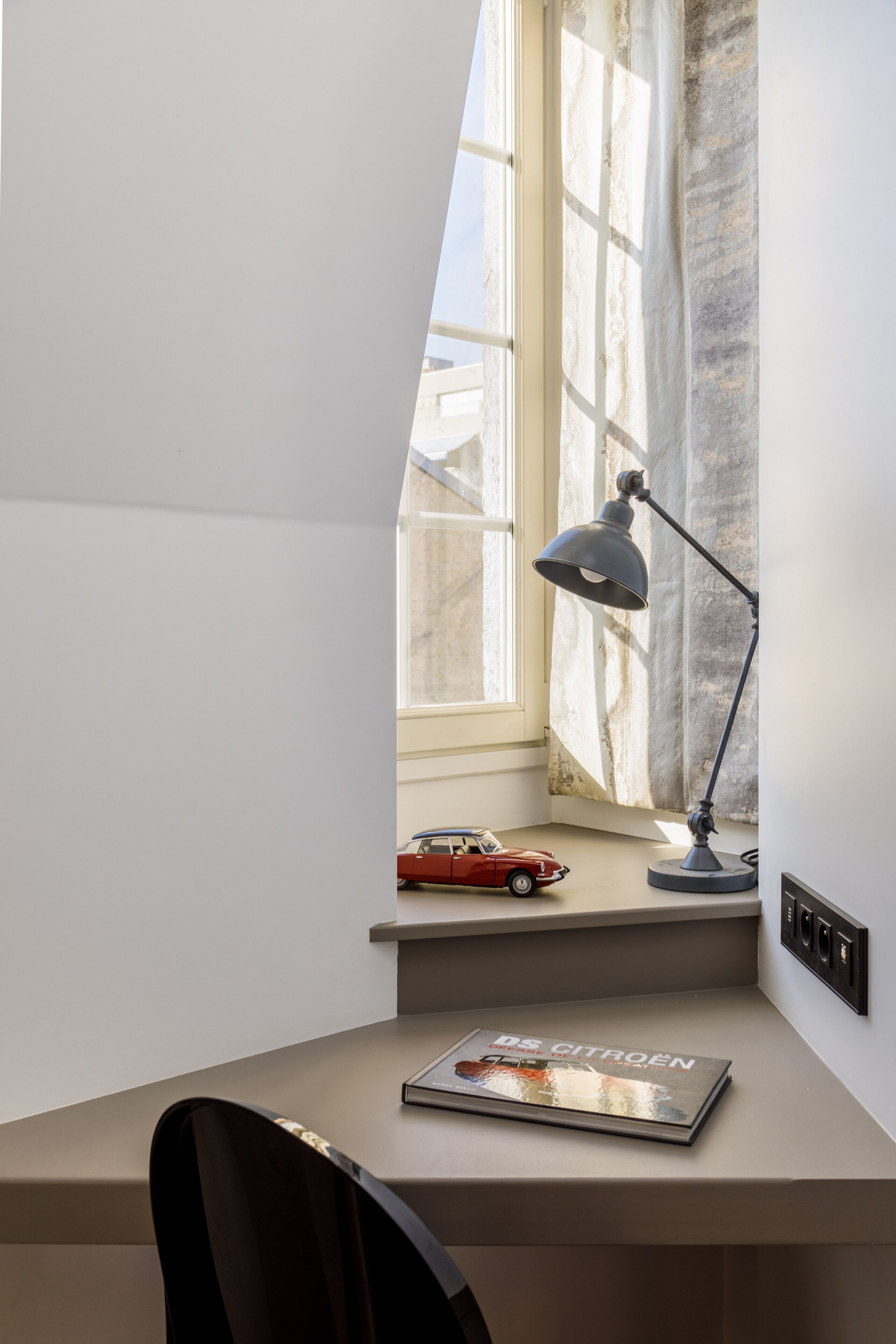 A desk with a lamp, a magazine, and a toy car by a window.