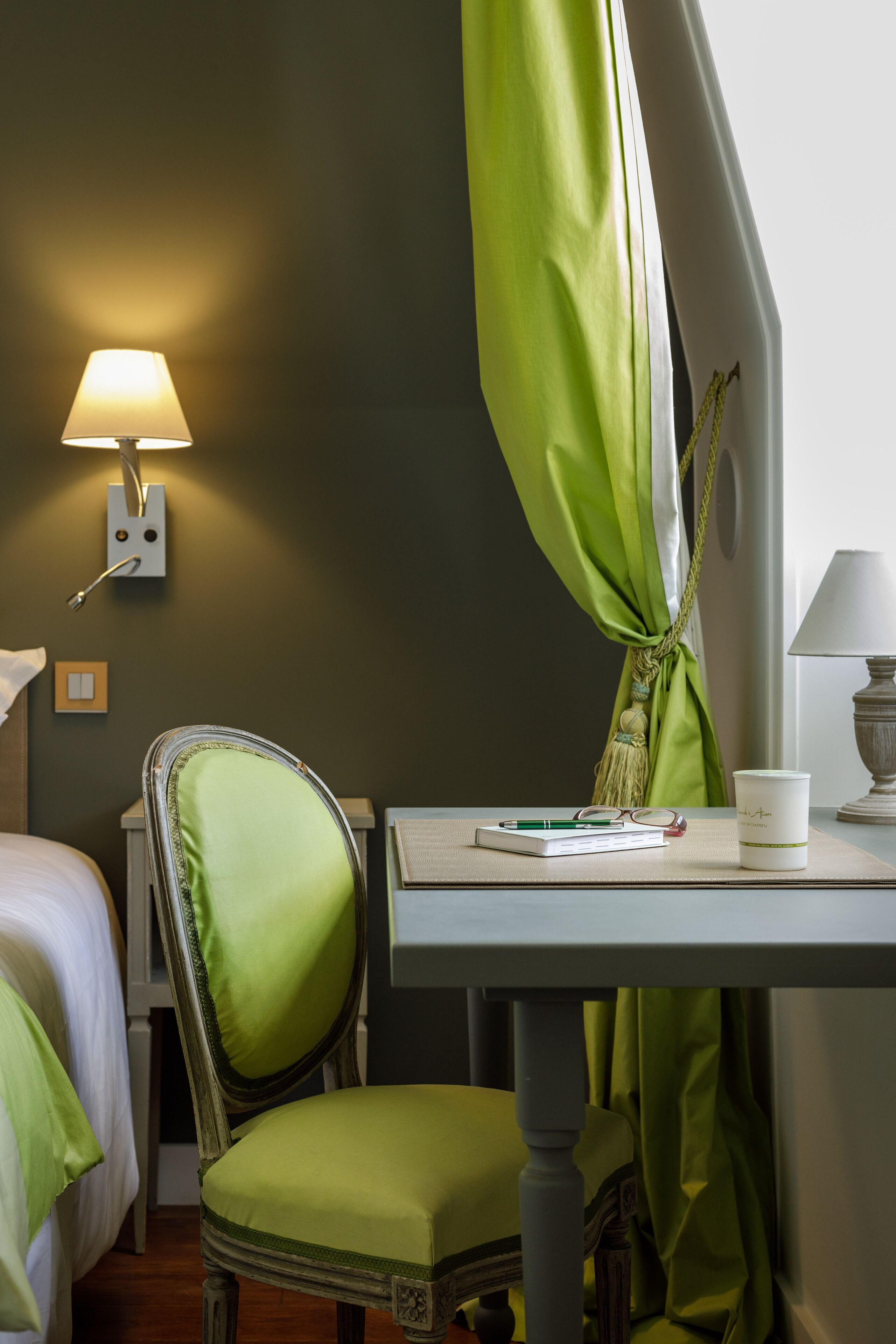 A hotel room with a bed, a desk with a lamp, a chair, and a window with green curtains.