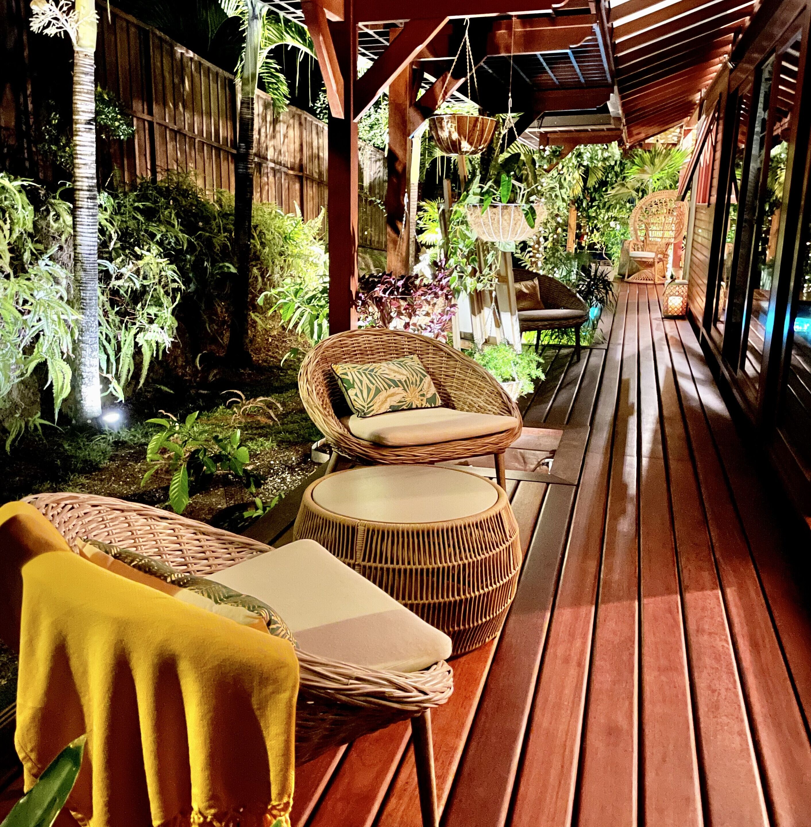 A wooden deck with wicker furniture, including chairs and a round table, surrounded by lush greenery and a wooden fence.