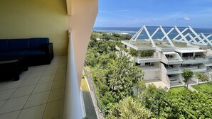 Un balcón con un sofá azul, vistas a un edificio moderno con una estructura de techo singular y un paisaje verde exuberante con árboles y un cielo azul despejado.