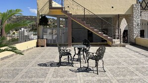 Un bâtiment doté d’un escalier menant à un balcon, d’un espace extérieur aménagé avec des chaises et une table en métal, et offrant une vue sur un paysage vallonné.