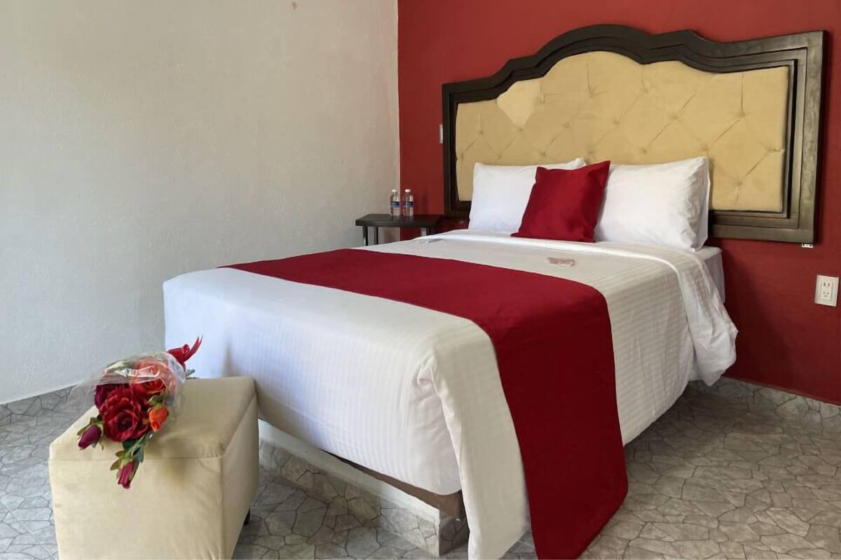 A hotel room with a bed, a nightstand, a small table, and a chair with a bouquet of flowers.