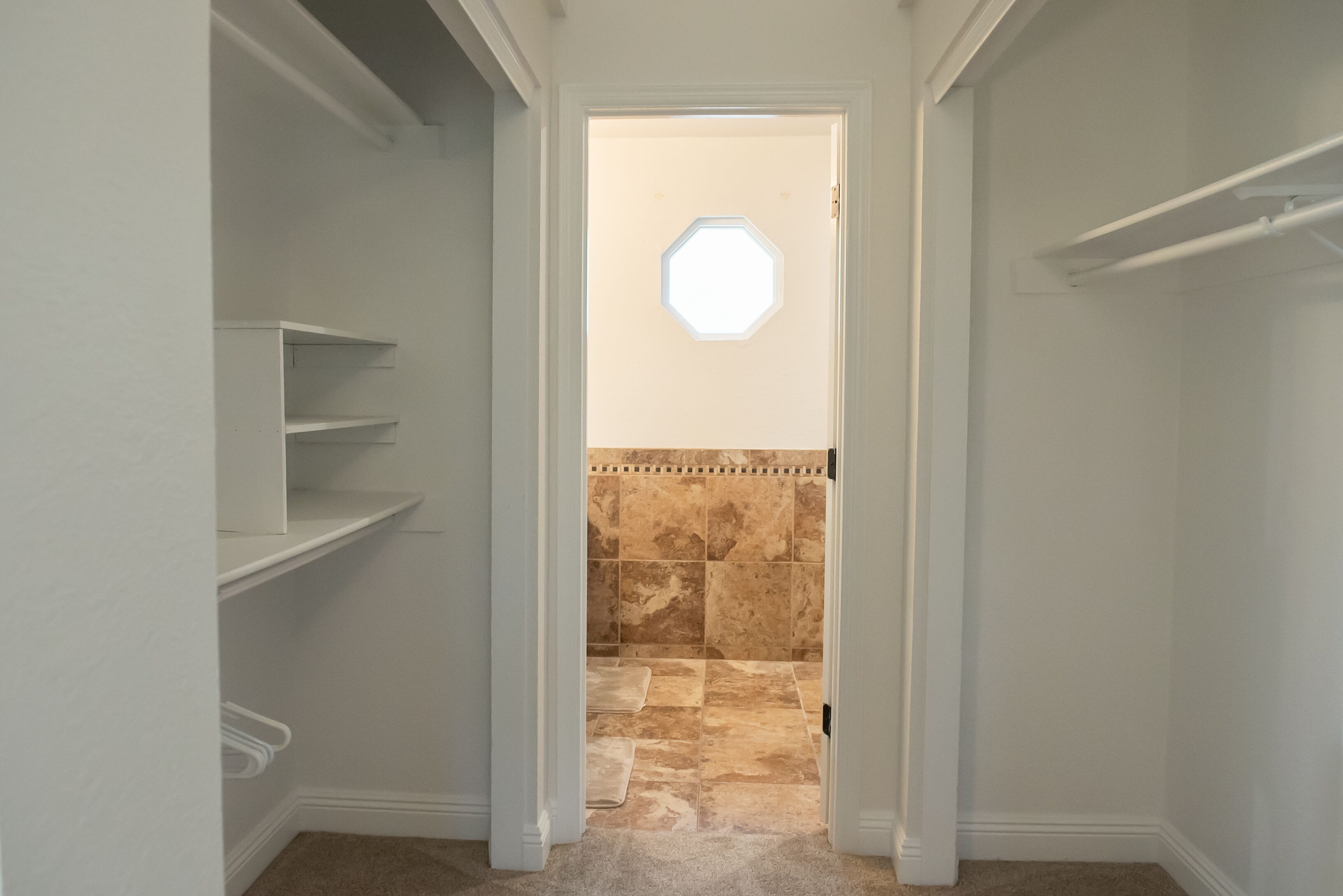 A narrow hallway with white walls, a beige carpet, and a tiled shower area with a small window.