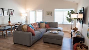 A modern living room with a grey sectional sofa, a small ottoman, a dining table with chairs, and a TV on a stand.