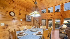A wooden cabin interior with a dining table set for four, wooden chairs, a chandelier, and a staircase leading to a balcony.