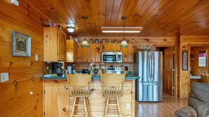 A wooden cabin interior with a kitchen featuring wooden cabinets, a stainless steel refrigerator, and two bar stools.