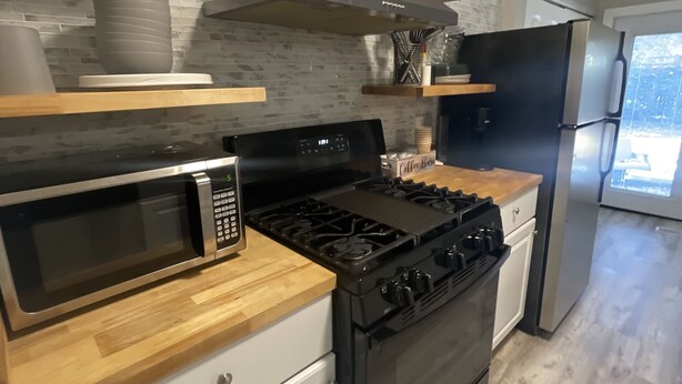 A modern kitchen with a stainless steel stove, microwave, and black refrigerator. There are wooden shelves above the counter and a large window providing natural light. - ***Southern Home *** (Atlanta)