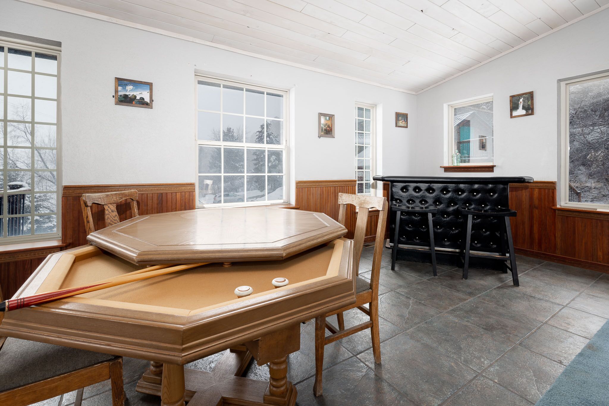 A billiards table with chairs, a pool table, and a bar area in a room with large windows and wooden paneling.
