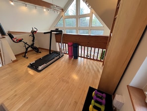 Een ruime kamer met houten vloer, een loopband, fitnessapparatuur en een groot raam met uitzicht op groen.