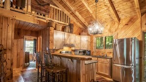 A rustic wooden cabin with a kitchen featuring a large island, bar stools, a refrigerator, and a hanging light fixture. - Luxury Cabin on the Lake & In The Trees (Saranac Lake)