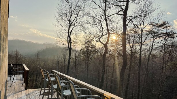 A wooden deck with metal chairs and a wooden railing overlooking a forested area with a view of the sun setting behind trees. - Windrock Retreat (Oliver Springs)