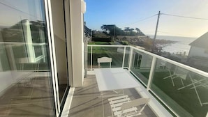 Un balcon doté d’une balustrade en verre, d’une chaise blanche et offrant une vue sur l’océan et les collines.