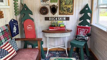 Une pièce chaleureuse avec un mur en bois, un canapé rouge, un fauteuil vert, une petite table avec un plateau, et divers éléments de décoration murale, notamment des bois de cerf et une enseigne sur laquelle on peut lire « BUCKSKIN ».