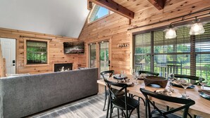 A cozy wooden cabin interior with a dining area, a sofa, and large windows offering a view of the outdoors. - Gorgeous Views! Secluded | HotTub | Quiet & Cozy (Townsend)