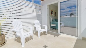 A balcony with two white plastic chairs, a potted plant, and a sliding glass door leading to a room with a blue couch and a small table. - Casa Felicidad, Air Conditioning, Sea View - Puerto del Carmen, Lanzarote (Puerto del Carmen)