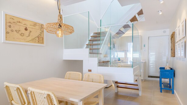 A modern interior with a dining area featuring a wooden table and wicker chairs, a staircase with glass railings, and a blue side table. - Casa Felicidad, Air Conditioning, Sea View - Puerto del Carmen, Lanzarote (Puerto del Carmen)