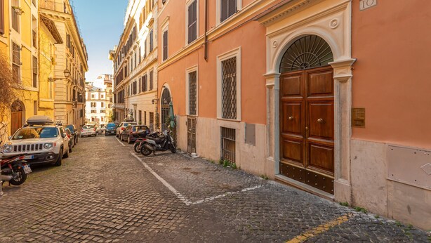 Une rue pavée bordée de voitures et de motos garées, des bâtiments à l’architecture classique et une porte en bois imposante sur la droite.