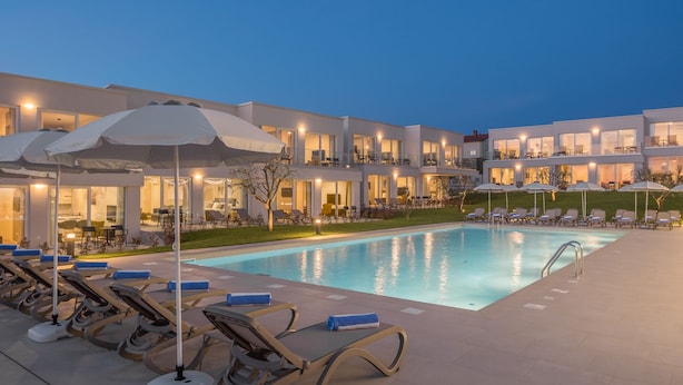 Um hotel moderno com piscina, espreguiçadeiras, guarda-sóis e fachada bem iluminada.