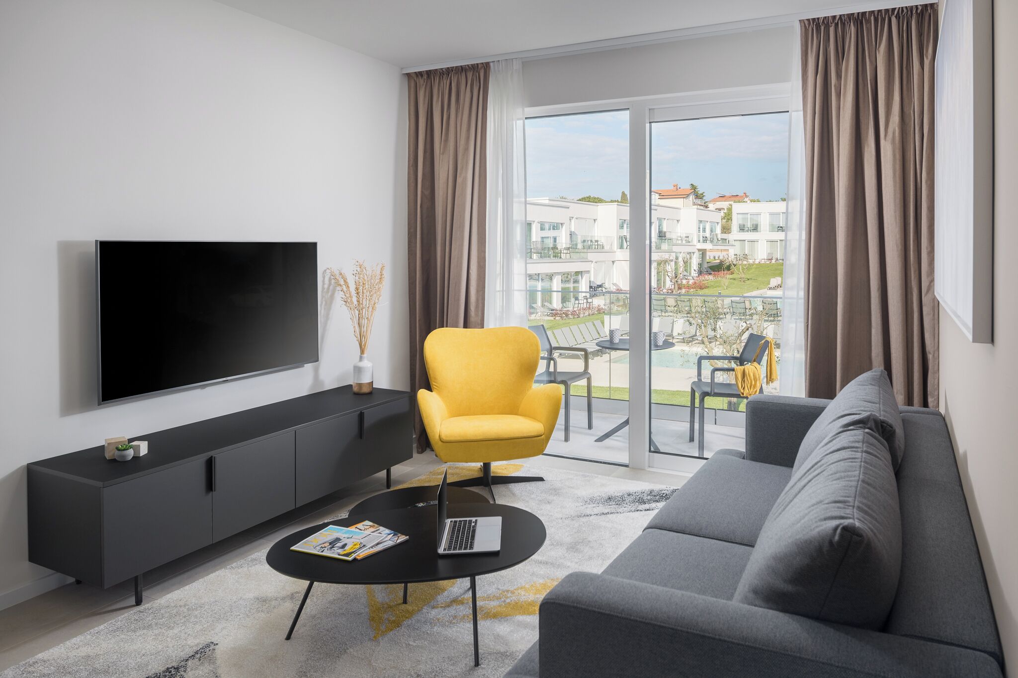Una sala de estar moderna con un televisor de pantalla plana, un sofá, un sillón amarillo y un balcón con vista a edificios.