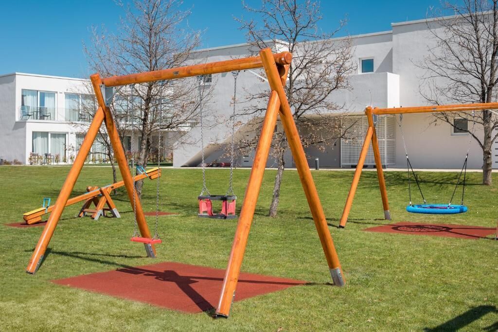 A playground with swings and a slide in a grassy area with a building in the background.