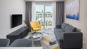 Uma sala de estar moderna com um sofá cinzento, uma poltrona amarela, uma televisão de tela plana e uma grande janela com vista para uma piscina e edifícios.