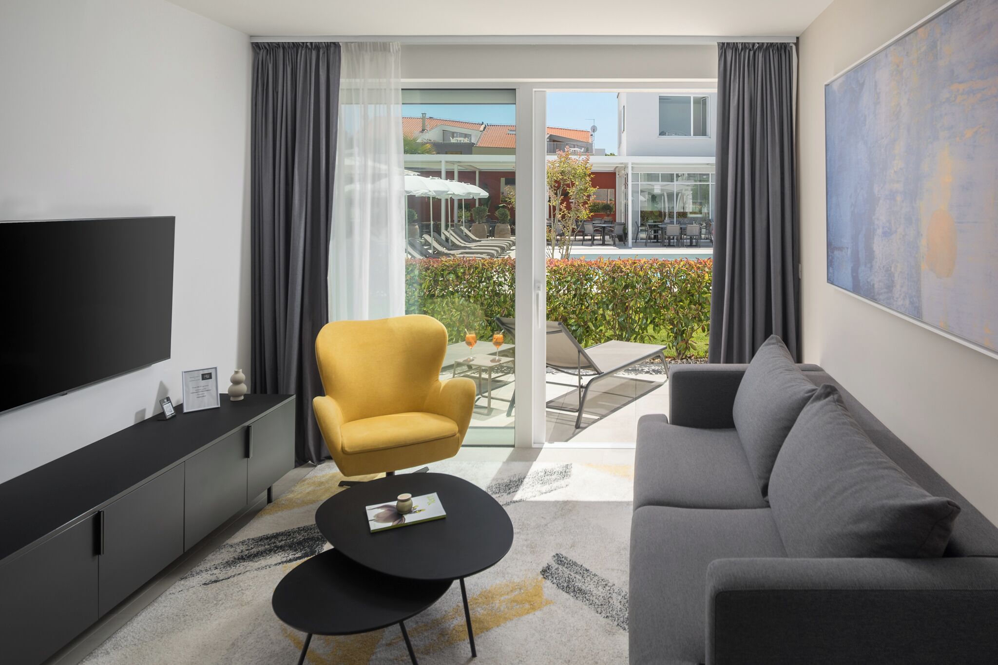 A modern living room with a flat-screen TV, a sofa, a yellow armchair, a small black table, and a glass door leading to an outdoor patio with a table and chairs.