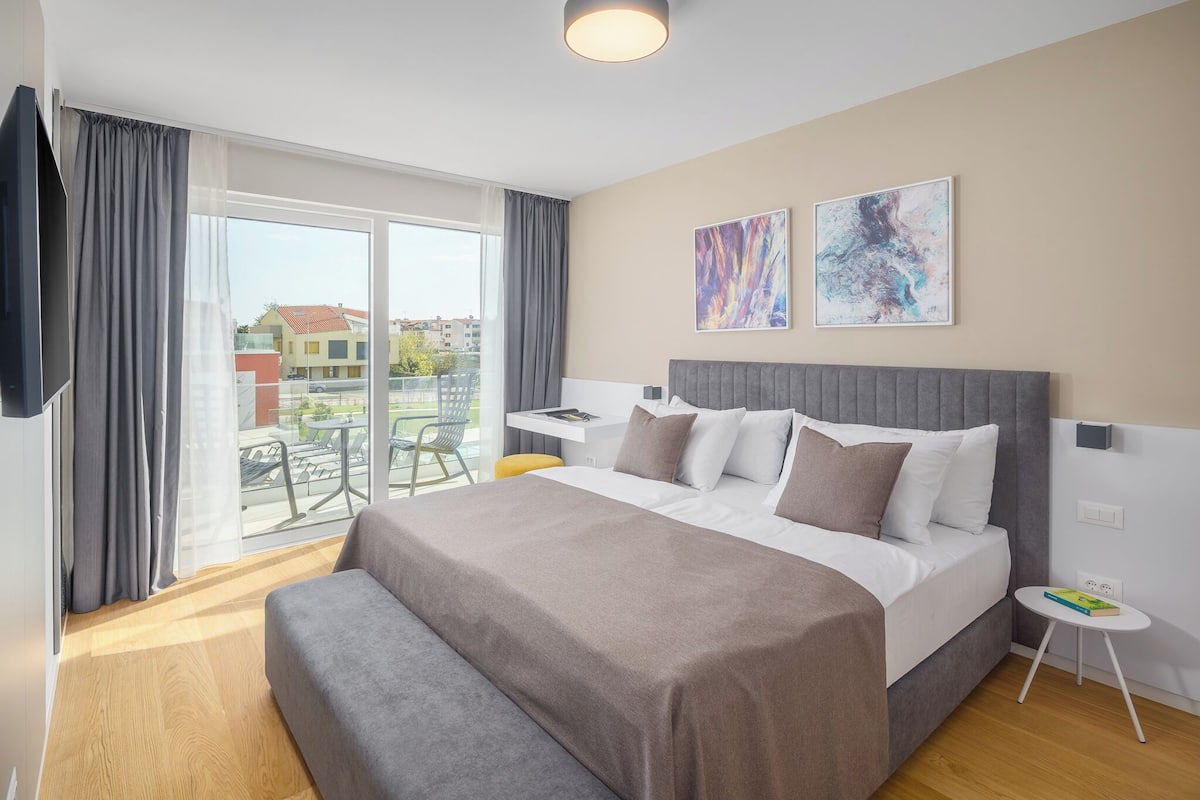 A modern hotel room with a large bed, a desk, and a balcony with outdoor seating.