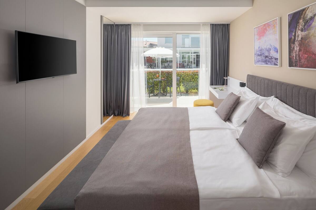 A modern hotel room with a large bed, a flat-screen TV, a balcony with an umbrella, and artwork on the walls.