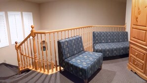 Una habitación de esquina con escalera de madera, dos sillones estampados, un armario de madera y una ventana con persianas.