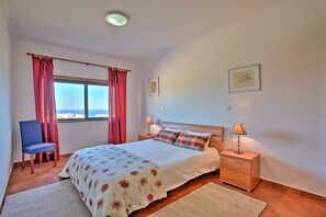 A bedroom with a bed, two bedside tables, a chair, a window with curtains, and a view of the sea.