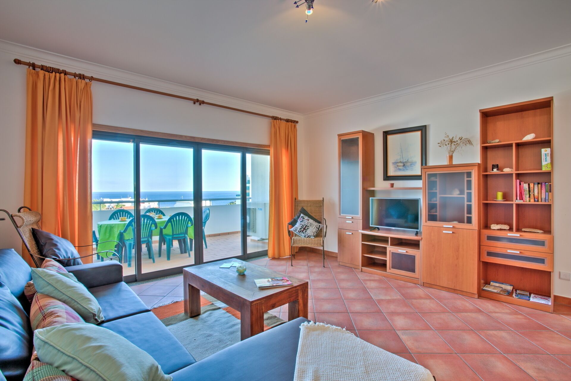 Een woonkamer met een bank, salontafel en tv-meubel. Er is een balkon met oranje gordijnen en uitzicht op de oceaan.