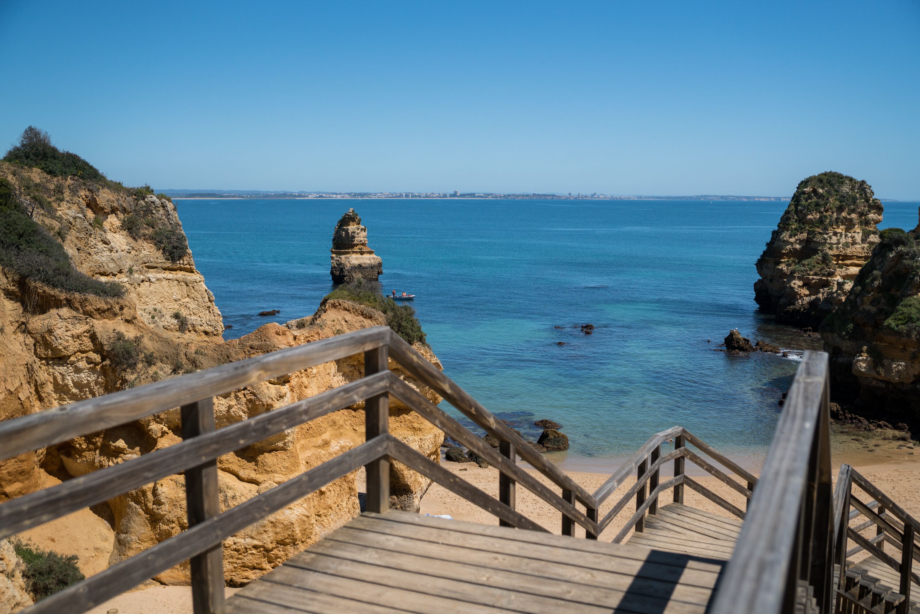 Eine Holztreppe führt zu einem Strand mit klarem, blauem Wasser und felsigen Klippen.