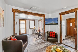 Un salón amplio con una mesa de comedor y sillas, un sofá con un cojín a cuadros rojo y negro, y piso de madera.