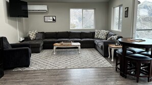 Een moderne woonkamer met een hoekbank, een salontafel, een televisie en een eethoek met barkrukken.