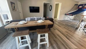 A dining area with a wooden table, two stools, and a black couch. A TV is mounted on the wall above the couch.