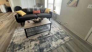 A modern living room with a black sofa, a coffee table, and a rug. There are large windows with blinds, a wall-mounted artwork, and a kitchen area in the background.