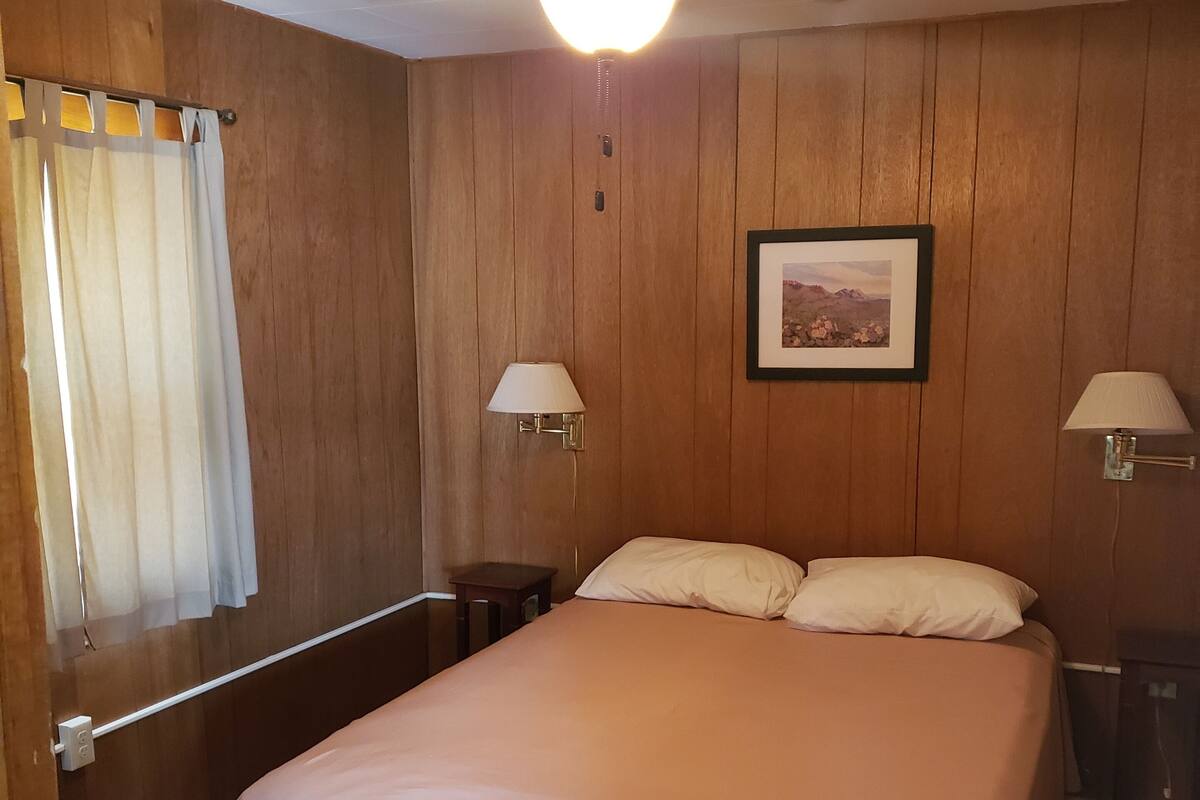 A bedroom with a bed, wooden walls, a ceiling fan, two wall-mounted lamps, and a framed picture on the wall.