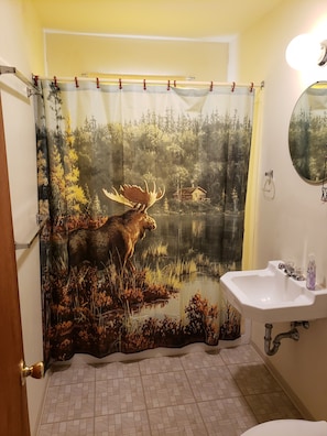 Un baño con cortina de ducha que muestra un alce en un bosque, un lavamanos y un espejo.