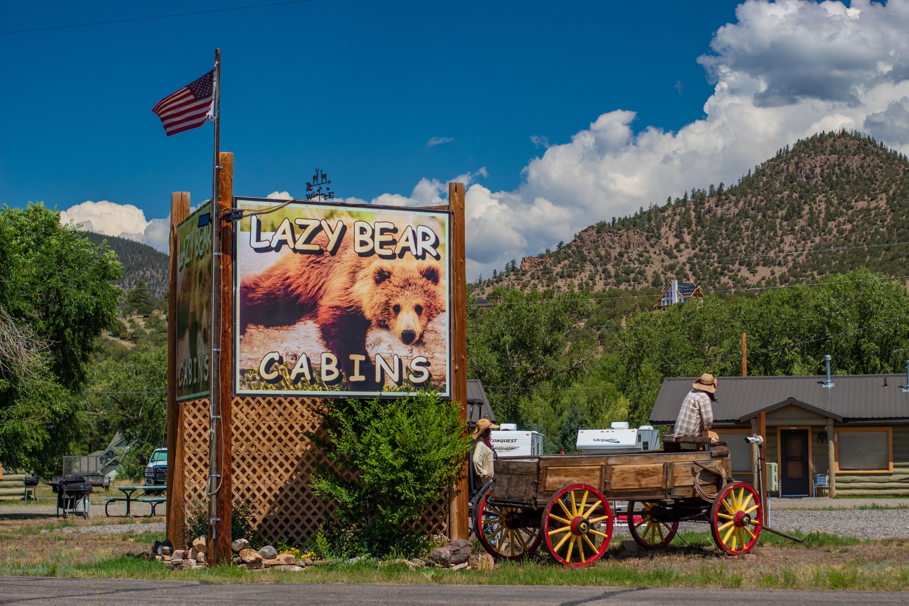 A sign for 'Lazy Bear Cabins' with a bear illustration, a wooden fence, and a wooden cart with red wheels.