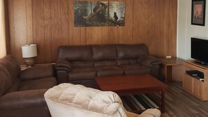 Ett vardagsrum med en hörnsoffa, en beige fåtölj, ett soffbord i trä, en tv och en takfläkt.