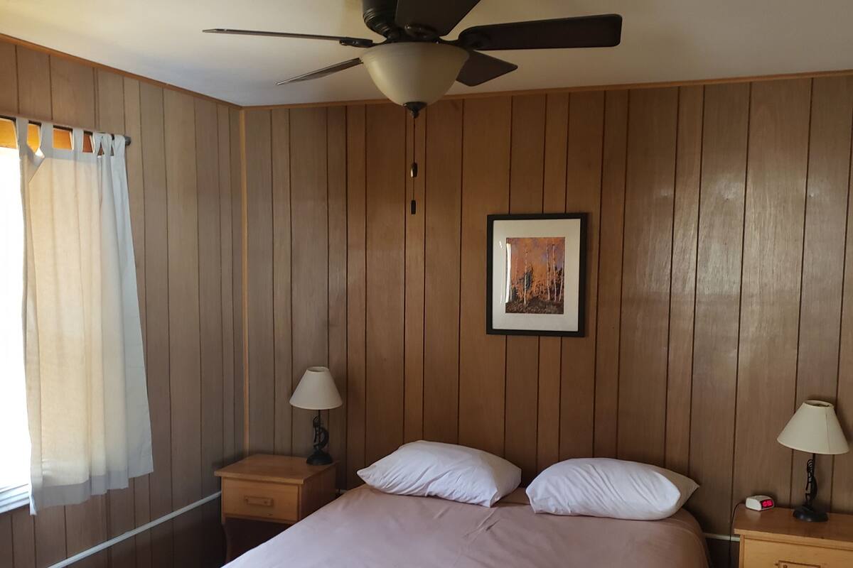 A bedroom with a bed, two bedside tables with lamps, a ceiling fan, wooden panel walls, a framed picture on the wall, and a window with curtains.