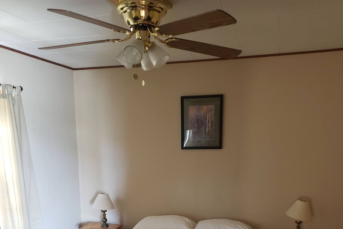 A bedroom with a bed, two bedside tables with lamps, a ceiling fan, a window with curtains, and a framed picture on the wall.