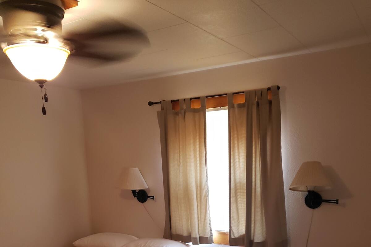 A room with two single beds, a small round table, a ceiling fan, and two wall-mounted lamps.