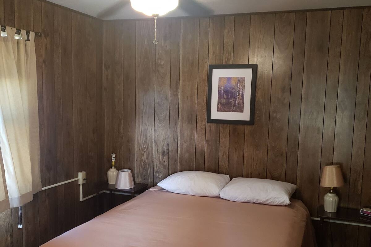 A bedroom with a wooden paneled wall, a ceiling fan, a framed picture, a bed with white linens, and bedside tables with lamps.