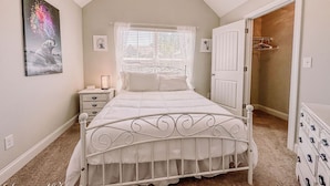 Een slaapkamer met een bed met wit metalen frame, een nachtkastje met een lamp, een raam met dunne gordijnen en een deur naar een kast.