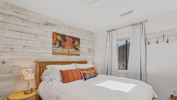 Ein Bett mit weißer Bettwäsche, einem hölzernen Kopfteil und dekorativen Kissen. Ein Nachttisch mit Lampe steht neben dem Bett. Ein Fenster mit Vorhängen ist an der Wand.