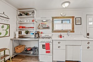 A compact kitchen with white cabinets, a stove, and a sink. There are shelves with various kitchen items and a window with a curtain.